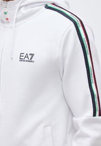 Bela jopa s kapuco na zadrgo z logotipom EA7 Emporio Armani na prsih in barvami italijanske zastave vzdolž črtastega traku na rami.
