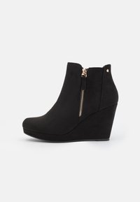 Černé kotníkové boty na wedge podpatku vyrobené z semiše, se bočním zipem a zlatými detaily. Design zahrnuje hladkou texturu a zaoblenou špičku.