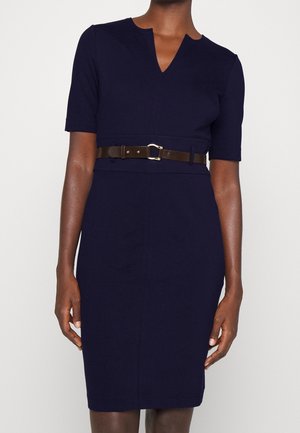 Frau trägt ein figurbetontes marineblaues Kleid mit halblangen Ärmeln und einem braunen Ledergürtel mit goldfarbener Schnalle in der Taille.