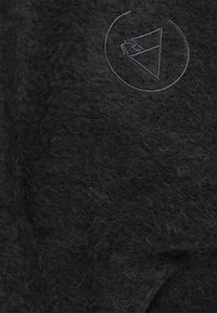 Zwarte pluizige stof met een gestikte driehoekige berg- en maanlogo binnen een cirkelvormige omlijning nabij de rechterbovenhoek.