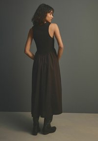 Zwart mouwloos jurkje met een aansluitend lijfje en een vloeiende rok, gemaakt van gladde stof. Model gestyled met zwarte laarzen tot de knie.