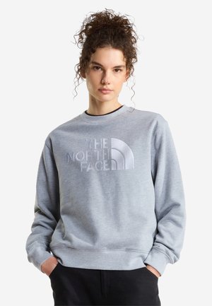 Nuori nainen, jolla on kiharat hiukset, pukeutuneena vaaleanharmaaseen collegeseen, jossa "The North Face" -logo, kädet mustien housujen taskuissa, neutraali tausta.