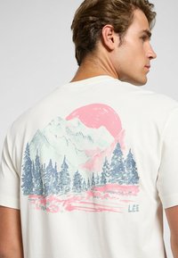 Camiseta de manga corta beige con una ilustración colorida de un paisaje natural que muestra montañas, pinos y un gran sol rosa en la parte posterior.