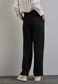Pantaloni neri con texture, dal taglio rilassato, con vita alta e tasche laterali. Abbinati a calzature di colore chiaro.