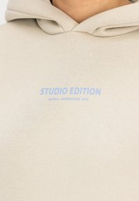 Beige hoodie van zachte stof, met een voorzak en een capuchon met trekkoord. Tekst "STUDIO EDITION" in lichtblauwe opdruk op de borst.