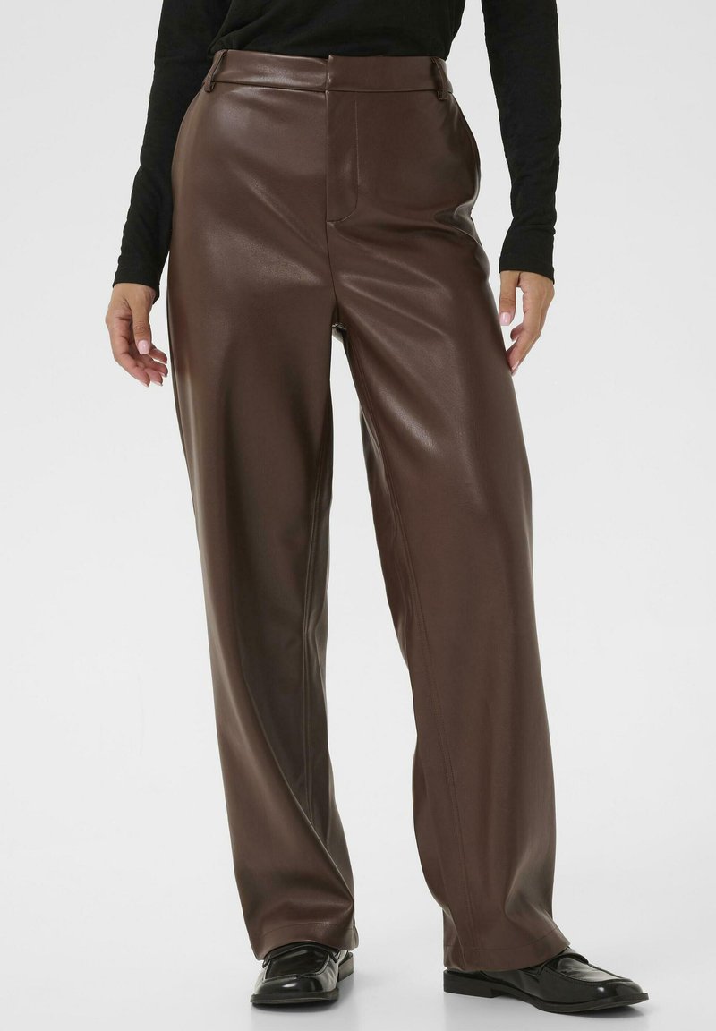 Personne portant un pantalon en cuir marron taille haute et jambes droites, un haut noir à manches longues et des mocassins noirs sur un fond blanc uni.