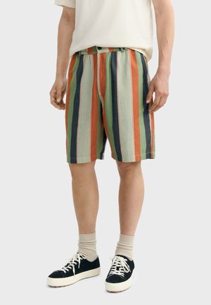 Persona con pantalones cortos hasta la rodilla con rayas verticales en verde, naranja, azul y beige, combinados con calcetas beige y zapatillas negras.