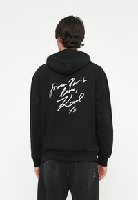 Felpa nera con cappuccio regolabile, con la scritta in bianco corsivo "da Parigi con amore, Karl" sul retro. Tessuto comodo, vestibilità rilassata.