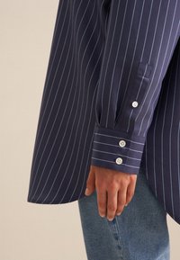 Camicia a righe blu navy con strisce verticali bianche, polsini doppi con tre bottoni bianchi e orlo curvo.
