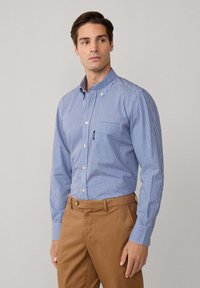 Man in een blauw-wit geruit overhemd met lange mouwen, gestoken in een tankleurige broek, staand tegen een effen lichte achtergrond, blikkend opzij.