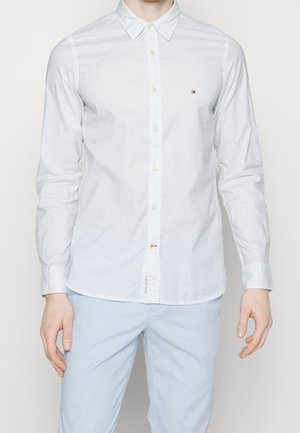 Mand iført en hvid langærmet skjorte med små prikker og lyseblå bukser, stående foran en ensfarvet baggrund.