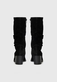 Bottes noires en daim jusqu'au genou avec un design froncé et une fermeture éclair sur le côté. Comprend un talon épais pour la stabilité et une surface texturée.