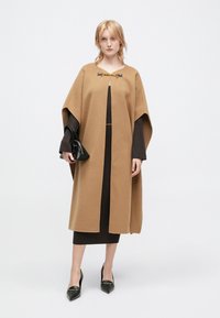 Καφέ κάπα από μείγμα μαλλιού με στρογγυλή λαιμόκοψη, διαθέτει μεταλλικό κούμπωμα. Φοριέται πάνω από ένα σκούρο, εφαρμοστό φόρεμα και μαύρη τσάντα.