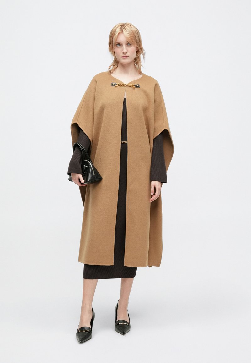 Καφέ κάπα από μείγμα μαλλιού με στρογγυλή λαιμόκοψη, διαθέτει μεταλλικό κούμπωμα. Φοριέται πάνω από ένα σκούρο, εφαρμοστό φόρεμα και μαύρη τσάντα.