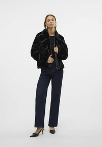 Czarna kurtka z sztucznego futra z kołnierzem, zamkiem błyskawicznym i krojem oversize, połączona z ciemnymi spodniami z szerokimi nogawkami i czarnymi sandałami na obcasie.