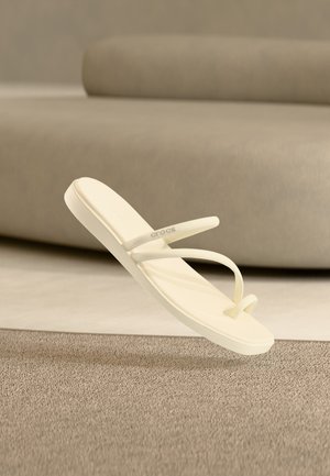 Smėlio spalvos minimalistinės basutės su plonais dirželiais ir piršto kilpa, pakibusios virš tekstūruoto kilimo neutralių tonų kambaryje su užapvalinta sofa fone.