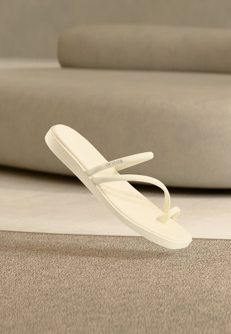 Μπεζ μινιμαλιστικό σανδάλι με δάχτυλο-λουράκι και λεπτές τιράντες, αιωρείται πάνω από υφασμένο χαλί σε δωμάτιο ουδέτερων τόνων με ένα καμπύλο καναπέ στο φόντο.