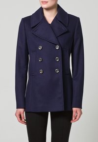 Tmavě modrý vlněný kabát typu pea coat s dvojitým zapínáním, velkými klopami a stříbrnými knoflíky. Má boční kapsy a vypasovaný střih.