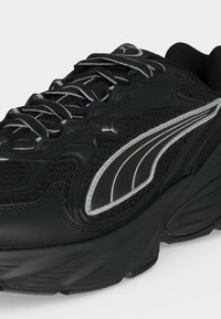 Μαύρο αθλητικό παπούτσι Puma με χοντρή σόλα, υφασμάτινες λεπτομέρειες, ανακλαστικό λευκό λογότυπο στο πλάι και μαύρα κορδόνια με λευκό περίγραμμα.