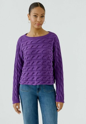 Femme portant des boucles d'oreilles créoles, un pull violet texturé et un jean bleu, debout devant un fond clair uni.