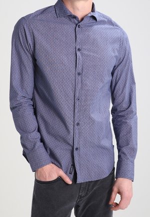 Man draagt een nauwsluitend blauw overhemd met knopen en een kleine stippenpatroon, gecombineerd met zwarte spijkerbroek, hand in de zak, tegen een effen achtergrond.