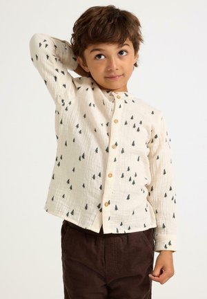Camicia button-up in tessuto crema con motivi neri a forma di albero; presenta un colletto alla cinese e bottoni in legno; abbinata a pantaloni marroni scuri.