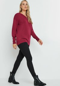Burgunderfarbener Oversized-Pullover mit V-Ausschnitt, kombiniert mit schwarzen eng anliegenden Leggings und Stiefeln mit Knöchelriemen. Glatte Textur, lässiges Design.