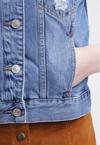 Blå denimjacka med metallknappar och slitna detaljer, hand instoppad i fickan på en brun kordflorskjol.