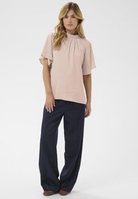 Blouse rose pâle avec des détails plissés et des manches volantes, associée à un pantalon large bleu marine et des chaussures tressées marron.