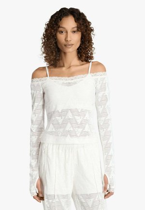 Donna con capelli ricci che indossa un top bianco in pizzo con spalle scoperte, decorato con motivi geometrici, e pantaloni larghi abbinati, su sfondo uniforme.