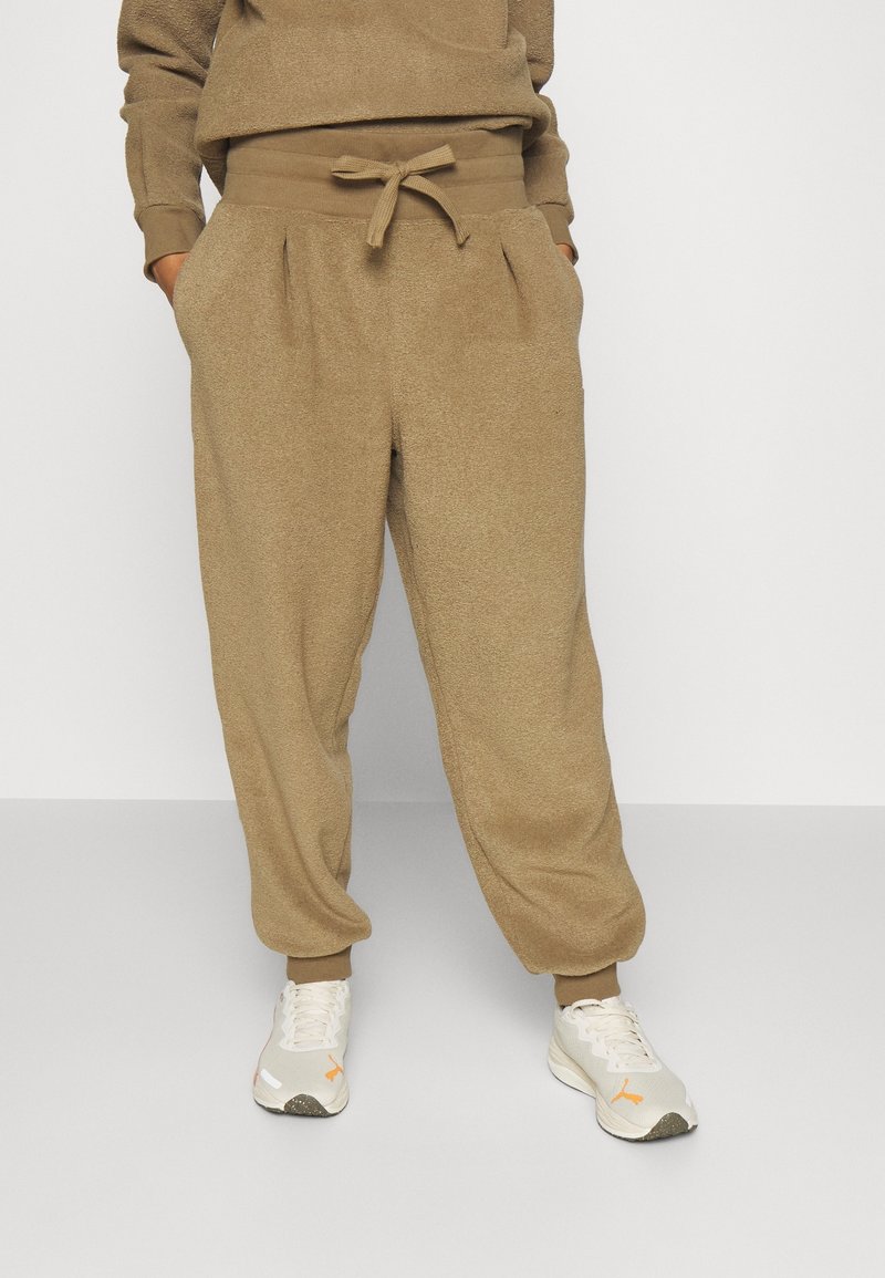 Personne portant un pantalon de jogging beige texturé avec une taille à cordon et des chaussures de sport blanches, debout devant un fond uni.