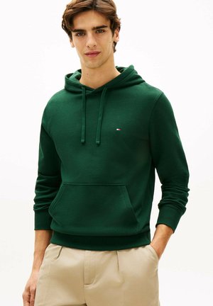 Giovane uomo che indossa una felpa con cappuccio verde scuro con tasca frontale e pantaloni beige, in piedi con una mano in tasca contro uno sfondo bianco.