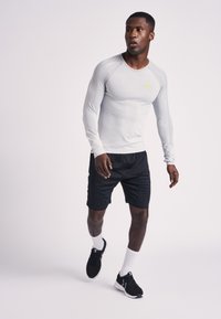 Maglietta sportiva a maniche lunghe grigio chiaro con trama a coste, logo giallo; abbinata a pantaloncini neri e scarpe da corsa nere, indossata con calzini bianchi.