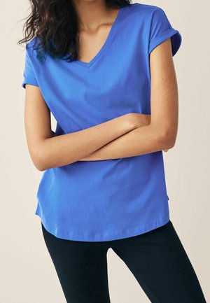 Vrouw met een blauw T-shirt met korte mouwen, V-hals en opgerolde mouwen, en een zwarte broek, met de armen over de borst gekruist.