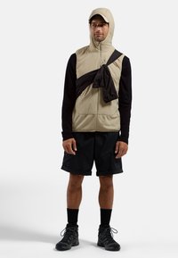 Beige sleeveless vest with hood, front zip, and patch pockets. Black fabric tied diagonally across the chest. Black shorts and hiking boots.