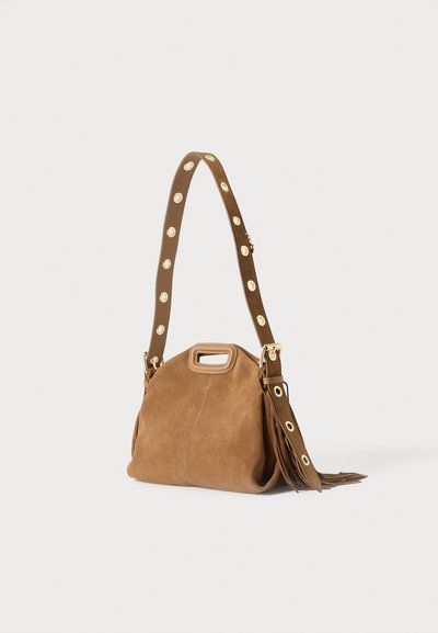 Sac à bandoulière en daim marron avec poignée découpée, sangle réglable ornée d'œillets dorés et détail pompon sur fond uni.