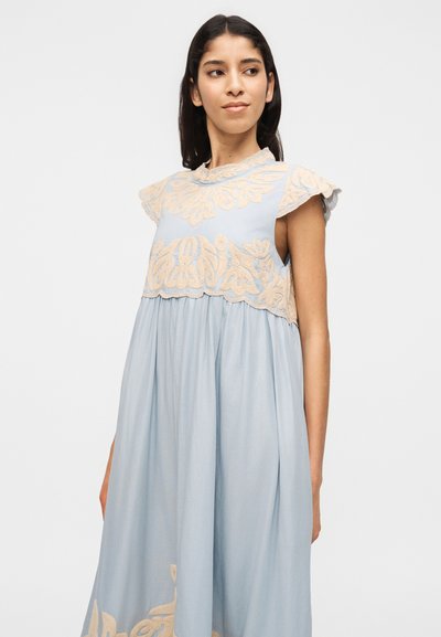 Γυναίκα με μακριά σκούρα μαλλιά που φοράει αμάνικο ανοιχτό μπλε φόρεμα με μπεζ κεντήματα λουλουδιών στο μπούστο και στο κάτω μέρος, κοιτάζοντας ελαφρώς προς τα δεξιά.