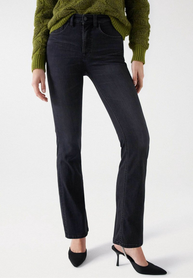 Femme portant un jean noir taille haute à jambes droites, un pull vert texturé et des chaussures à talons noires à bout pointu avec des brides à la cheville.