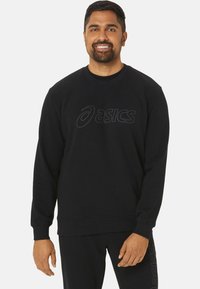 Sweatshirt noir à col rond, avec un logo texturé sur la poitrine. Le tissu semble doux avec des poignets et un ourlet côtelés.
