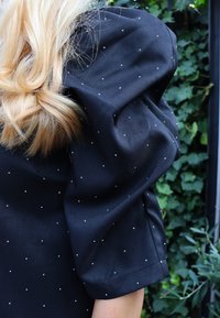 Bluză din material negru cu mâneci bufante, cu mici buline albe. Textură netedă cu design structurat și cusături vizibile.