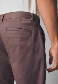 Brązowe bawełniane spodnie o gładkiej fakturze, z tylną kieszenią o minimalistycznym designie i dopasowanym kroju, prezentowane na dolnej części pleców.