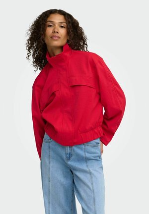 Femme aux cheveux bouclés portant une veste rouge à col montant et un jean bleu clair, debout devant un fond uni.