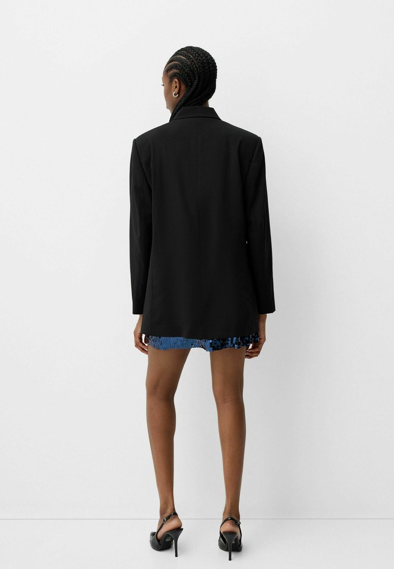 Blazer noir avec épaules structurées, présentant un ourlet bleu pailleté. Tissu lisse, manches longues et longueur sous les hanches.
