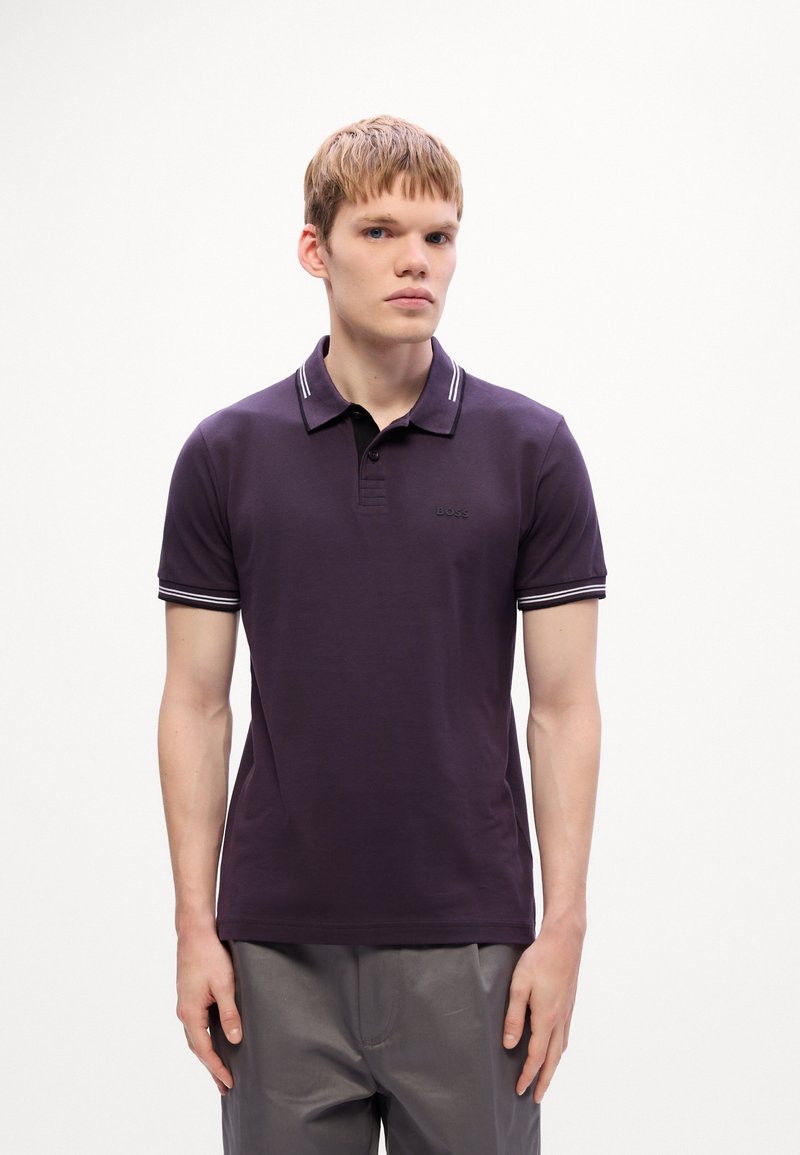 Jeune homme portant un polo violet foncé avec un col et des poignets rayés, se tenant devant un fond blanc uni.