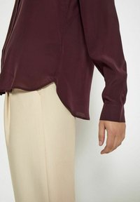 Blusa borgogna fluida in tessuto traslucido, con maniche lunghe e orlo curvo. Abbinata a pantaloni strutturati beige chiaro.