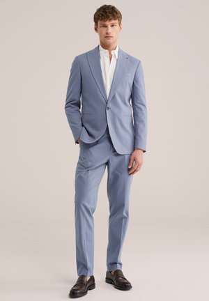 Uomo con abito azzurro chiaro, camicia bianca e mocassini marrone scuro, in piedi con una mano in tasca davanti a uno sfondo semplice.