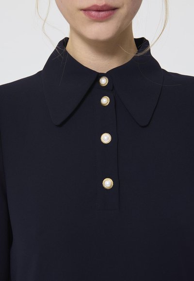 Sinine kraejuhtmega blouse, millel on viis kuldse aktsendiga nuppu, mille keskel on pärlid, valminud sujuvast kangast ja puhtast disainist.