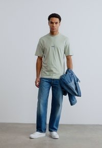 Άνδρας με ανοιχτό πράσινο μπλουζάκι και μπλε τζιν, κρατώντας ένα διπλωμένο μπλε ριγέ μπουφάν, στέκεται μπροστά σε λευκό απλό τοίχο.