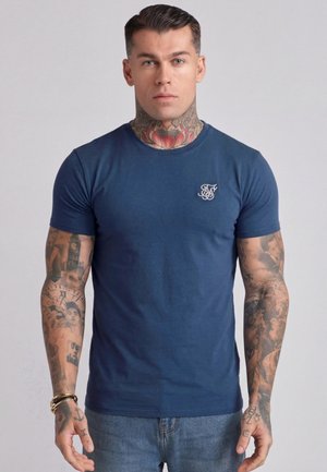 Mann mit kurzen dunklen Haaren, umfangreichen Tätowierungen an Hals und Armen, trägt ein eng anliegendes dunkelblaues T-Shirt und blaue Jeans, steht vor einem einfarbigen Hintergrund.