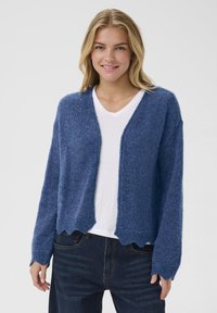 Blue knitted cardigan features a loose fit, scalloped hem, and textured fabric. Worn over a white V-neck shirt.